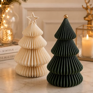 Könnte beinhalten: Zwei dekorative Weihnachtsbaum-Ornamente mit einer gefalteten, papierartigen Textur. Ein Baum ist cremefarben mit einem Stern als Spitze, der andere ist dunkelgrün mit einer kleinen goldenen Kugel an der Spitze. Beide haben eine konische Form.
