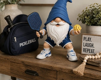 May include: A garden gnome figurine wearing a blue hat, blue shirt, and blue shorts, holding a blue pickleball paddle and a yellow pickleball. The gnome wears white athletic sneakers with blue accents. A navy blue bag with the word PICKLEBALL is nearby.