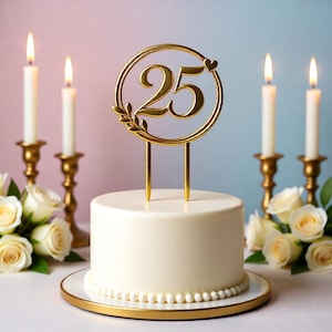 May include: A white cake with a gold rimmed plate and a gold "25" cake topper. The topper is in a circle with a heart and leaf design. Lit candles in gold holders and white roses are in the background.