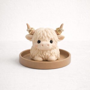 May include: A beige Highland cow figurine with a round tray base. The cow has fluffy fur, small horns, and rings on its horns. The figurine is a decorative item for holding rings or small jewelry.