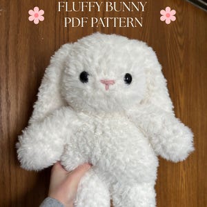 May include: A fluffy white bunny plush toy with black eyes and a pink nose. The bunny has long, floppy ears and is made of a soft, textured material. The text "FLUFFY BUNNY PDF PATTERN" is displayed at the top.