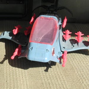 May include: A vintage toy set featuring a purple action figure, a gray and blue helicopter with red accents, a missile, and a small silver gun. The action figure is standing next to a card that reads "SEA SLUG".