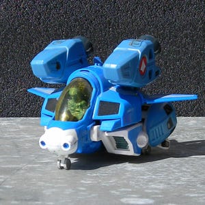 May include: A blue and white toy aircraft with a clear cockpit and a green figure inside. The toy has two large blue engines on top and small wings. The aircraft is on a gray surface.