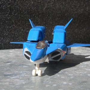 Puede incluir: Un avión de juguete azul y blanco con una cabina transparente y tren de aterrizaje. El juguete tiene dos motores grandes con detalles circulares en blanco y negro. Las alas y los estabilizadores son azules. El juguete está sobre una superficie gris.