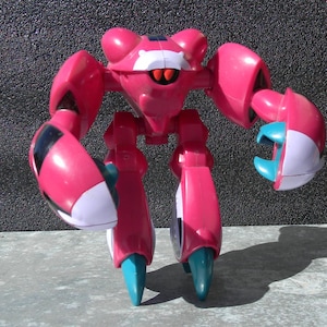 Puede incluir: Un robot de juguete rosa y blanco con un diseño similar a un cangrejo. El robot tiene brazos grandes, piernas y una cabeza con ojos rojos. Las garras y los pies del robot son de color azul claro. El juguete está hecho de plástico y está sobre una superficie gris.