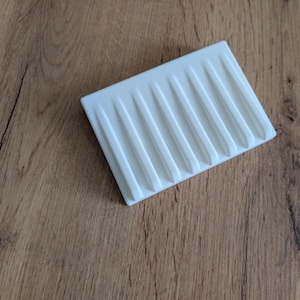 Può includere: Un portasapone rettangolare bianco con un design a coste. Il portasapone è realizzato in un materiale solido e si trova su una superficie di legno. Le scanalature parallele corrono lungo la lunghezza del portasapone, offrendo funzionalità e decorazione.