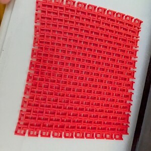 May include: A vibrant red, interlocking plastic mesh sheet. The sheet is composed of numerous small, square-shaped openings, creating a grid-like pattern. The material appears to be flexible and lightweight, suitable for various applications.