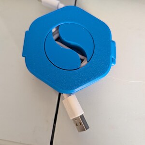 以下が含まれることがあります： 白のUSBケーブルと白のUSB-Cケーブルを備えた、青色の六角形のケーブルオーガナイザー。オーガナイザーは、ケーブルを収納するための中央の湾曲したスロットを備えたユニークなデザインです。ケーブルは白です。