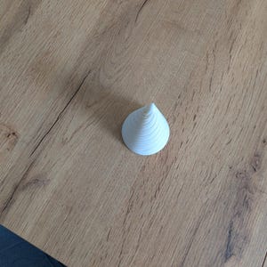 May include: A small, white, conical object with a spiral design, resting on a light brown wooden surface. The object has a pointed top and a rounded base, with visible concentric rings. The wooden surface has a natural wood grain pattern.