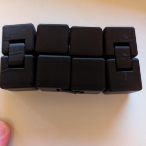May include: A black fidget toy with a series of connected, rectangular blocks. The blocks are hinged, allowing them to bend and move. The toy is designed to be manipulated and played with, offering a tactile experience.