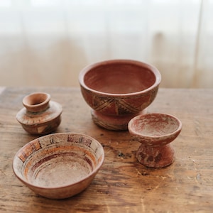 May include: Four terracotta pottery pieces, including bowls and a small vase, are arranged on a wooden surface. The bowls feature geometric patterns in shades of brown and orange. The pottery has a rustic, handmade appearance.