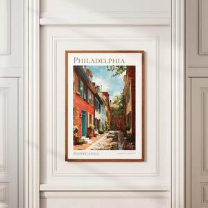 May include: A framed art print of a Philadelphia street scene. The print features colorful row houses in red, blue, and white, with a cobblestone street. The word "PHILADELPHIA" is at the top, and "PENNSYLVANIA" is at the bottom.
