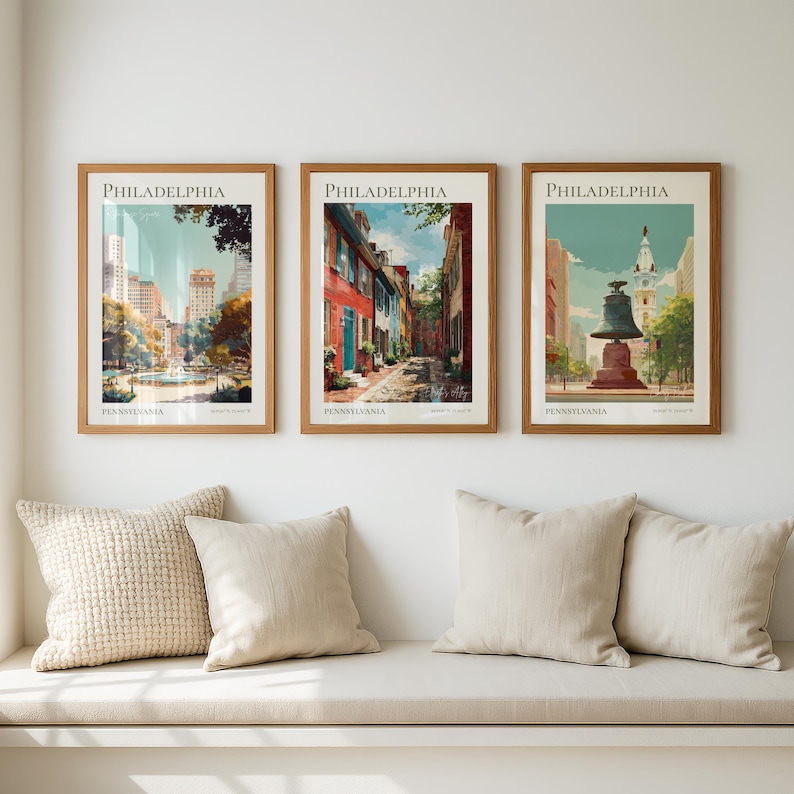 May include: Three framed Philadelphia art prints with a vintage aesthetic. Each print features a different city scene, including a cityscape, a historic street, and the Liberty Bell. The prints are in wooden frames and are displayed above a bench with pillows.