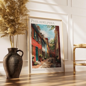 May include: A framed art print of a Philadelphia street scene. The print features colorful buildings with red, blue, and white facades. The artwork is displayed on a wooden floor, next to a decorative vase with dried plants.