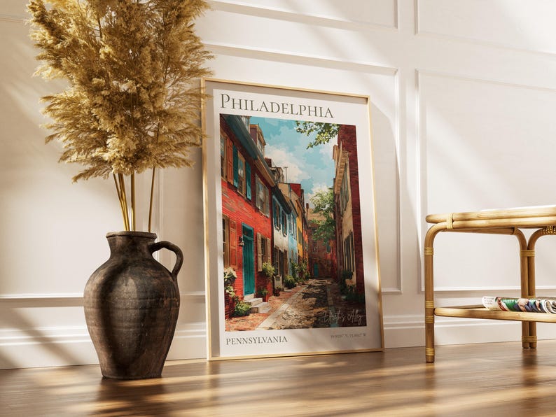 May include: A framed art print of a Philadelphia street scene. The print features colorful buildings with red, blue, and white facades. The artwork is displayed on a wooden floor, next to a decorative vase with dried plants.