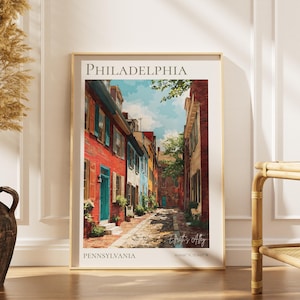 May include: A framed art print featuring a colorful street scene in Philadelphia. The artwork depicts a narrow cobblestone alley lined with brick buildings in shades of red, blue, and beige. The print is labeled "PHILADELPHIA" and "PENNSYLVANIA".