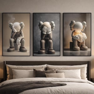 May include: Three framed art prints depicting a seated cartoon figure in different poses. The figure is mainly beige with black details, and each print has a different background colour. The prints are displayed above a bed with neutral-toned bedding.