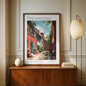 May include: Framed art print of Elfreth's Alley in Philadelphia, Pennsylvania. The artwork depicts a colorful street scene with red, blue, and yellow buildings. The text "PHILADELPHIA" and "PENNSYLVANIA" are at the top and bottom of the print.