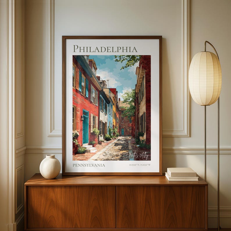 May include: Framed art print of Elfreth's Alley in Philadelphia, Pennsylvania. The artwork depicts a colorful street scene with red, blue, and yellow buildings. The text "PHILADELPHIA" and "PENNSYLVANIA" are at the top and bottom of the print.