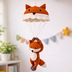Puede incluir: Un juguete de zorro tejido suspendido de un dosel en forma de paracaídas. El zorro es naranja, blanco y marrón, con una cara sonriente. El dosel es naranja y blanco, con un borde festoneado. Ideal para una habitación infantil.