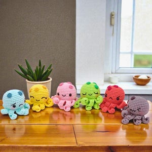 Könnte beinhalten: Sechs gehäkelte Oktopus-Spielzeuge in verschiedenen Farben: blau, gelb, rosa, grün, rot und lila. Jeder hat einen anderen Gesichtsausdruck. Sie sind auf einer Holzoberfläche angeordnet, mit einer Topfpflanze im Hintergrund.