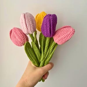 Könnte beinhalten: Ein Strauß handgehäkelter Tulpen in Rosa, Lila und Gelb, mit grünen Stielen und Blättern. Die Blumen werden zusammengehalten und zeigen die leuchtenden Farben und das strukturierte Garn, perfekt für eine Frühlingsdekoration.
