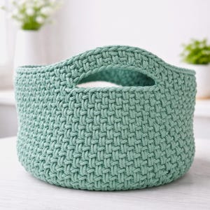 Könnte beinhalten: Ein runder, gewebter Korb in einem weichen, gedämpften Grünton. Der Korb hat einen stabilen Griff und eine strukturierte Oberfläche. Der Korb ist wahrscheinlich handgefertigt und eignet sich zur Aufbewahrung oder Dekoration.