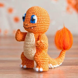 Könnte beinhalten: Gehäkelte orangefarbene Charmander-Figur mit cremefarbenem Bauch, blauen Augen und einem Flammenschwanz. Das handgefertigte Amigurumi-Spielzeug steht und hat den Daumen hoch. Die Figur ist ein Charakter aus dem Pokémon-Franchise.