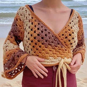 Könnte beinhalten: Ein gehäkeltes Wickeloberteil in Braun-, Beige- und Orangetönen. Es hat einen V-Ausschnitt, lange Ärmel mit ausgestellten Manschetten und einen Bindverschluss in der Taille. Das offene Webmuster ist ein Hauptmerkmal des Kleidungsstücks.