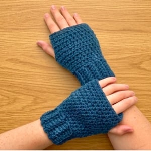 Könnte beinhalten: Ein Paar handgefertigte, tealblaue Fingerhandschuhe. Die Handschuhe sind mit einem strukturierten Stich gehäkelt und haben eine gerippte Manschette und offene Finger. Die Handschuhe sind so konzipiert, dass sie die Hände warm halten und gleichzeitig die Fingerfertigkeit ermöglichen.