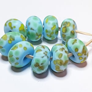 Può includere: Una collezione di perline di vetro fatte a mano nei toni dell'azzurro, del verde e del giallo. Le perline hanno un motivo maculato e sono infilate su una fibra naturale. Ideali per la creazione di gioielli.