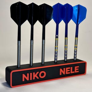 Könnte beinhalten: Ein Satz Darts mit schwarzen und blauen Flights, präsentiert in einem schwarz-roten Halter. Der Halter trägt den Namen "NIKO NELE" in roten Buchstaben. Die Darts haben silberne Schäfte.