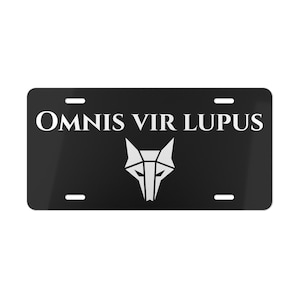 Op de afbeelding: Zwarte nummerplaat met de witte tekst "OMNIS VIR LUPUS" en een wit geometrisch wolfskopontwerp. De plaat heeft vier montagegaten en een glanzende afwerking.