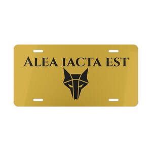 Op de afbeelding: Een goudkleurige nummerplaat met de zwarte tekst "ALEA IACTA EST". Daaronder staat een geometrisch wolfskopontwerp, eveneens in zwart. De plaat heeft vier montagegaten.