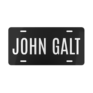 May include: A black license plate with the words "JOHN GALT" in bold, white capital letters. The plate has four small, white dashes near the top and bottom edges, and is rectangular with rounded corners.