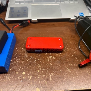 以下が含まれることがあります： 小さな画面が付いた赤い長方形の電子機器が木製の表面の中央に配置されています。黒い箱が左側の青い長方形のオブジェクトの上にあります。背景にはワイヤーとラップトップがあります。