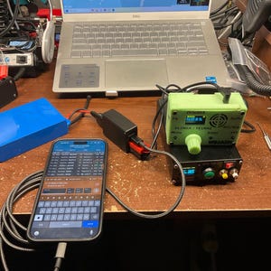 May include: A workspace with electronic devices, including a laptop, a smartphone displaying data, and a green and black electronic device. A blue rectangular battery pack and various cables are also present on the wooden surface.