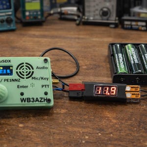 May include: A mint green (t)uSDX transceiver with a digital display showing radio frequency data, connected to a black battery holder with three green batteries. A digital voltage meter displays 8.8. The setup is on a wooden surface.