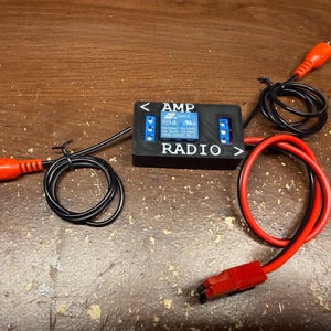 Peut inclure: Un composant électronique noir avec l'inscription "AMP RADIO", des connecteurs bleus et des fils rouges et noirs. Le composant comporte des connecteurs RCA rouges et un connecteur d'alimentation rouge. Les fils sont enroulés et connectés au composant.