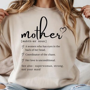 May include: Beige sweatshirt with the word "mother" in a cursive font, accompanied by a heart symbol. Below the word is a definition: "A women who has eyes in the back of her head. Coordinator of the chaos. Her love is unconditional. See also: superwoman, strong, not your maid."