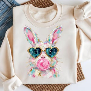 以下が含まれることがあります： ハート型のサングラスをかけたウサギが、ガムの泡を吹いているカラフルなイラストが描かれた、クリーム色のスウェットシャツ。スウェットシャツは、青いジーンズと茶色の織りのプレースマットと組み合わせています。
