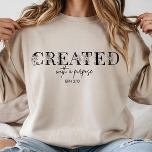 Puede incluir: Sudadera beige con la frase "CREATED with a purpose" en letras negras y detalles florales. El texto "EPH 2:10" está debajo. La sudadera tiene mangas largas y cuello redondo.