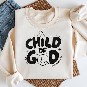Puede incluir: Sudadera color crema con el texto "CHILD OF GOD" en una fuente negra juguetona, con una corona y una carita sonriente. La sudadera se combina con vaqueros azules. El diseño incluye el versículo bíblico Gálatas 3:26.