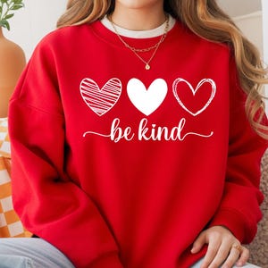 May include: A red sweatshirt with three white hearts and the words "be kind" in a cursive font. The sweatshirt is a solid red color and has a crew neck. The hearts are in different styles.