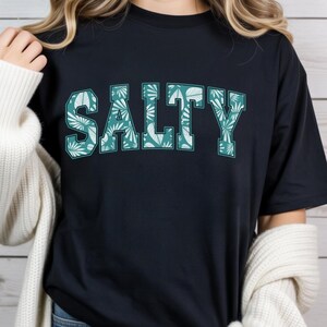 May include: A navy blue t-shirt with the word "SALTY" in large, teal block letters. The letters are filled with a tropical leaf pattern. The shirt has a crew neck and short sleeves. The overall style is casual and relaxed.