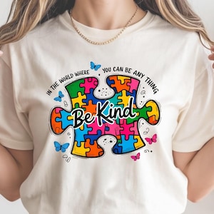 Puede incluir: Camiseta color crema con un diseño de pieza de rompecabezas colorido y las palabras "Be Kind" en escritura cursiva. El diseño incluye mariposas y la frase "In the world where you can be anything".
