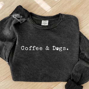 Op de afbeelding: Een donkergrijze sweatshirt met de witte tekst "Coffee & Dogs" en een pootafdruk. De longsleeve ligt op een lichtbruin houten oppervlak.