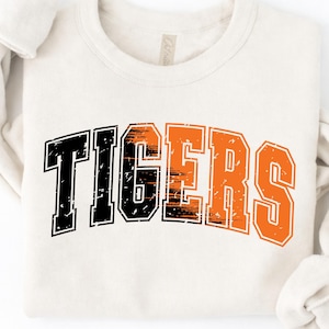 May include: A cream-colored long-sleeve sweatshirt featuring the word TIGERS printed in a distressed, arched varsity font. The letters transition from black on the left to a vibrant orange on the right, with a textured, weathered appearance.
