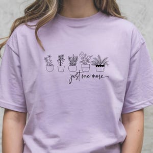 May include: A light purple t-shirt featuring a black line drawing of five potted plants. Below the plants, the script reads "just one more". The shirt is a solid color and has short sleeves.