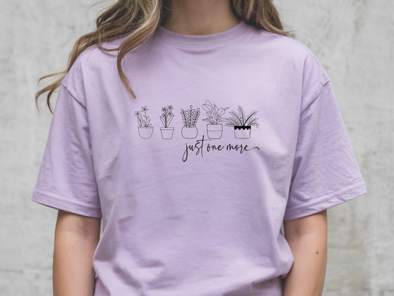 May include: A light purple t-shirt featuring a black line drawing of five potted plants. Below the plants, the script reads "just one more". The shirt is a solid color and has short sleeves.